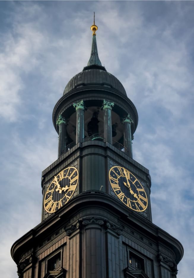 Nahaufnahme des oberen Teils des Hamburger Michel-Turms mit seinen markanten Uhren und der goldenen Spitze, vor einem bewölkten Himmel. Erinnert an die lange Tradition der PENSIONSKASSE der Hamburger Hochbahn Aktiengesellschaft -VVaG- in Hamburg